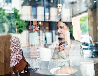 Durch das Fenster Blick auf fröhliche Millennial diverse Freundinnen plaudern glücklich, während an der Theke im Café sitzen und genießen heißen Kaffee mit Gebäck - ADSF18227