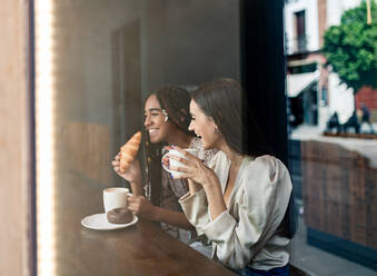 Durch das Fenster Blick auf fröhliche Millennial diverse Freundinnen plaudern glücklich, während an der Theke im Café sitzen und genießen heißen Kaffee mit Gebäck - ADSF18228