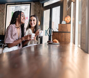 Side view of cheerful young multiracial women in trendy outfits drinking coffee and chatting happily while spending time together in modern cafeteria - ADSF18229