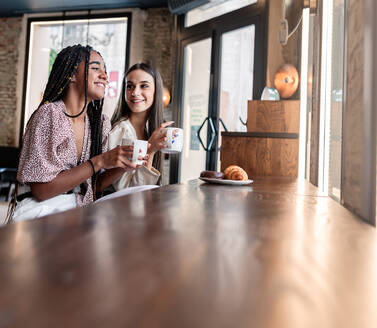 Seitenansicht von fröhlichen jungen multirassischen Frauen in trendigen Outfits, die Kaffee trinken und fröhlich plaudern, während sie in einer modernen Cafeteria Zeit miteinander verbringen - ADSF18229