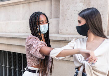 Side view of positive young multiracial female friends in trendy clothes and protective masks greeting each other with elbow bump on city street during coronavirus pandemic - ADSF18230