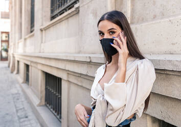 Junge Frau in modischer Kleidung und Schutzmaske, die in der Nähe eines steinernen Gebäudes in einer Stadt steht und sich während einer Coronavirus-Epidemie über ihr Smartphone unterhält und in die Kamera schaut - ADSF18232