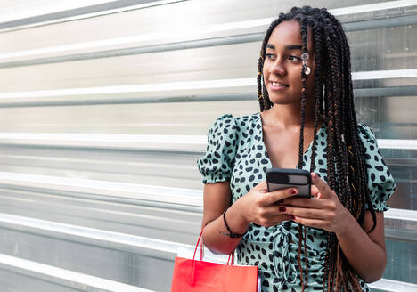 Inhalt Afroamerikanische Frau im Sommerkleid steht mit Einkaufstasche in der Stadt in der Nähe einer Metallwand und liest Nachrichten auf dem Handy - ADSF18236