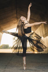 Elegant ballerina with eyes closed in black tutu and pointe shoes balancing on leg while moving gracefully during rehearsal in passage in city - ADSF18242