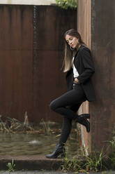 Thoughtful young female professional standing by pond while leaning on brown wall - MTBF00712