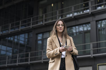 Smiling businesswoman holding mobile phone while standing against office building - MTBF00726