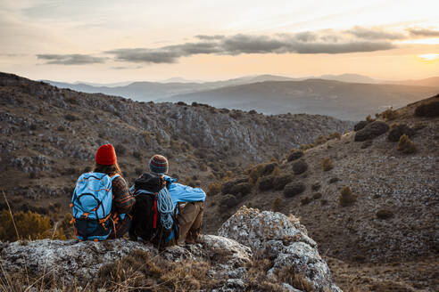 Male and female hikers enjoying sunset while sitting on rock mountain during vacation - RCPF00420