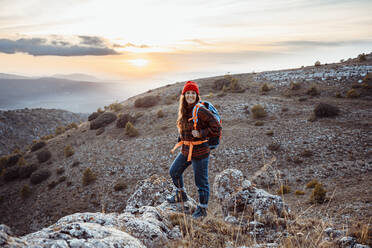 Lächelnde Wanderin, die wegschaut, während sie bei Sonnenuntergang auf einem Felsen steht - RCPF00421