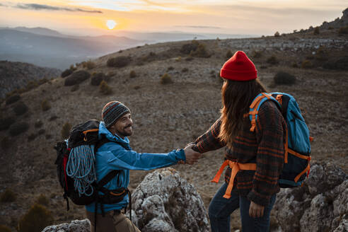 Girlfriend helping boyfriend to climb rocky mountain during sunset - RCPF00429