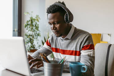 Männlicher Freiberufler mit Kopfhörern, der im Heimbüro einen Laptop benutzt - VABF04086
