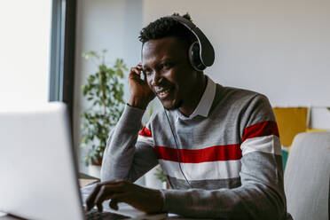 Lächelnder männlicher Unternehmer mit Kopfhörern, der zu Hause einen Laptop benutzt - VABF04087