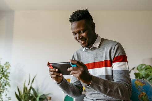 Lächelnder Geschäftsmann, der während seiner Arbeitspause zu Hause ein Handheld-Videospiel spielt - VABF04097