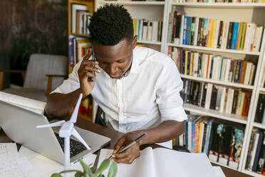 Männlicher Umweltschützer mit Windturbinenmodell, der in seinem Büro zu Hause mit seinem Smartphone telefoniert - VABF04102