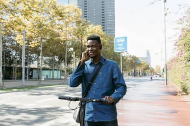 Männlicher Pendler, der mit einem elektrischen Roller auf dem Bürgersteig in der Stadt mit seinem Smartphone telefoniert - VABF04120