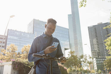 Male entrepreneur using smart phone with electric push scooter against buildings in city - VABF04121