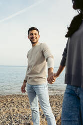 Smiling man holding woman's hand while standing at beach - VABF04141