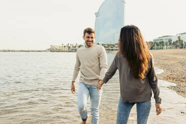 Smiling couple holding hands while walking on water at beach - VABF04143