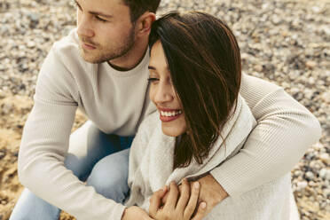 Young couple embracing each other while sitting on sand at beach - VABF04152