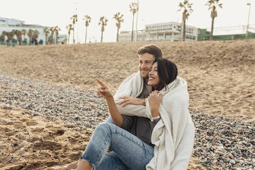 Smiling girlfriend pointing while embracing boyfriend at beach - VABF04153