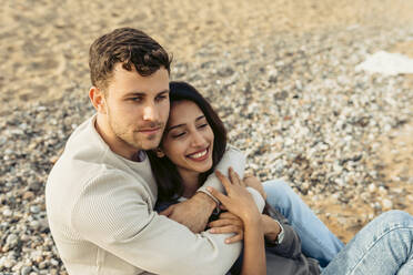 Young couple embracing while sitting on sand at beach - VABF04157