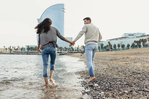 Freundin und Freund halten sich an den Händen, während sie am Strand übers Wasser laufen - VABF04161