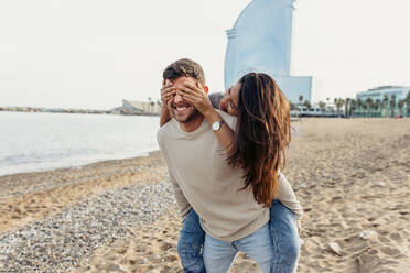 Freund nimmt ihn huckepack, während seine Freundin ihm am Strand die Augen zuhält - VABF04164