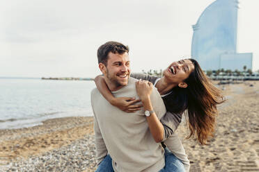 Ein lächelnder Mann nimmt eine fröhliche Frau am Strand huckepack - VABF04165