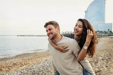 Freund lächelt, während er seine Freundin am Strand huckepack nimmt - VABF04166