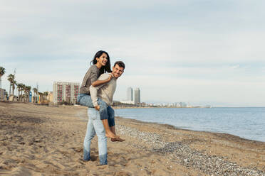 Young man giving piggyback ride to woman against sky at beach - VABF04167