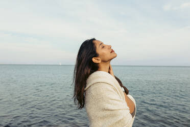 Young woman with eyes closed standing against sky at beach - VABF04176