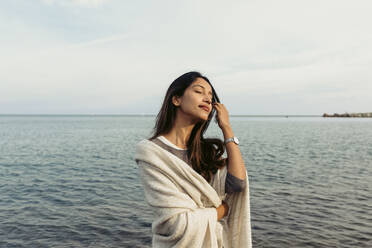 Woman with hand in hair standing against sea at beach - VABF04177