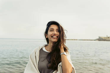 Lächelnde Frau mit Hand im Haar, die gegen den klaren Himmel am Strand wegschaut - VABF04182