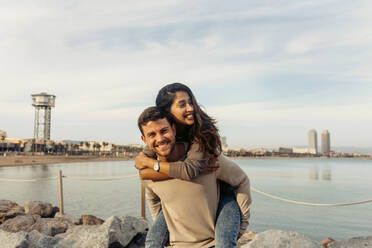 Smiling boyfriend giving piggyback ride to girlfriend against sea and sky - VABF04197