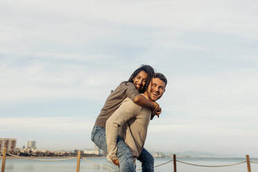 Happy boyfriend giving piggyback ride to girlfriend standing by sea against sky - VABF04198