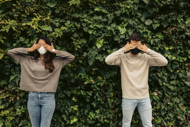 Junges Paar, das seine Augen mit den Händen vor Pflanzen im Park abdeckt, während COVID-19 - VABF04202