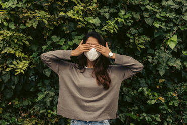 Young woman wearing protective face mask while covering eyes with hands against leaves in park - VABF04203