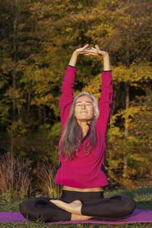 Reife Frau mit geschlossenen Augen hebt die Arme, während sie im Herbst im Park Yoga übt - FCF01935