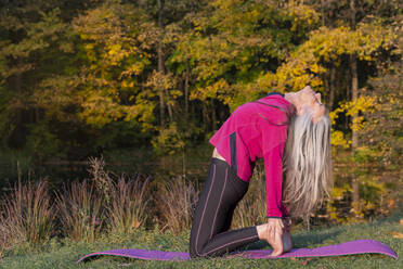 Mature woman stretching on yoga mat at park during autumn - FCF01936