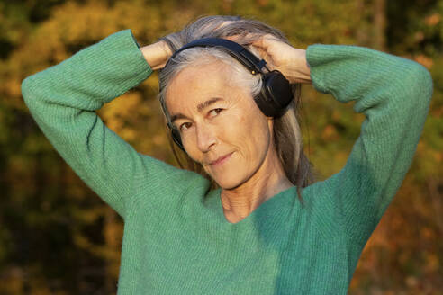 Lächelnde reife Frau hört Musik über Kopfhörer, während sie mit den Händen im Haar steht - FCF01939