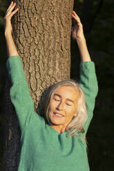 Mature woman with eyes closed against tree trunk at park during autumn - FCF01940