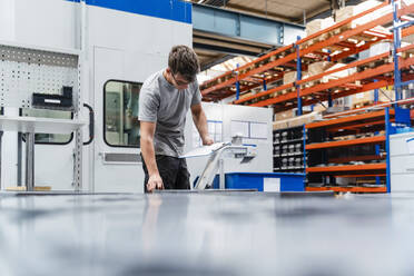 Junger männlicher Ingenieur, der ein Klemmbrett hält, während er eine Maschine in der Industrie untersucht - DIGF13432