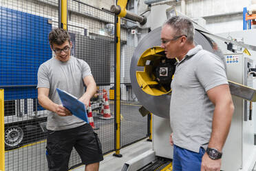 Smiling male colleague explaining mature man while standing by machinery at factory - DIGF13447