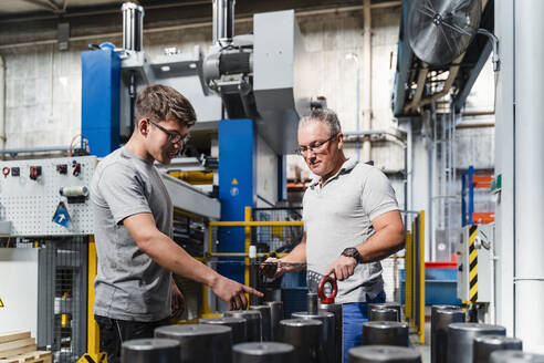 Junger männlicher Ingenieur im Gespräch mit einem reifen Mann über ein Produkt in einer beleuchteten Fabrik - DIGF13457