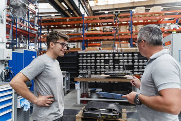 Lächelnder männlicher Mitarbeiter betrachtet einen reifen Ingenieur in einer beleuchteten Fabrik - DIGF13460