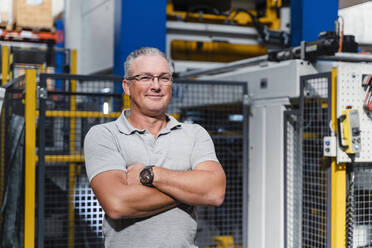 Confident male engineer with arms crossed standing in factory - DIGF13462