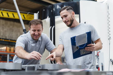 Älterer männlicher Techniker bei der Qualitätskontrolle eines Produkts, während er mit einem jungen Mitarbeiter in einer beleuchteten Fabrik steht - DIGF13469