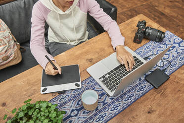 Von oben anonyme Fotografin, die ein Grafiktablett und einen Laptop benutzt, während sie am Tisch mit einer Fotokamera sitzt und an einem Projekt arbeitet - ADSF18301