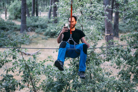 Excited young man photographing with mobile phone while enjoying zip line ride at adventure park stock photo