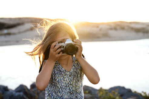 Unerkennbare junge Touristin in modischem Kleid, die das Meer am felsigen Ufer bei Sonnenuntergang fotografiert - ADSF18343