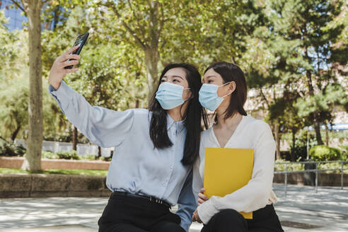 Unrecognizable young Asian female managers in classy clothes and medical masks relaxing in green park and taking selfie on smartphone - ADSF18354